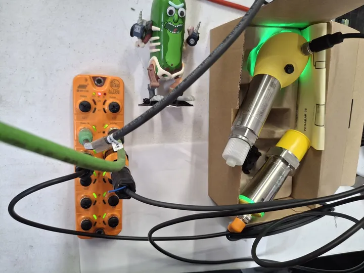 Bench test rig: an orange IO-Link master with lit status LEDs and networked cabling, alongside two stainless process transmitters, proving live data capture into WinCC Unified